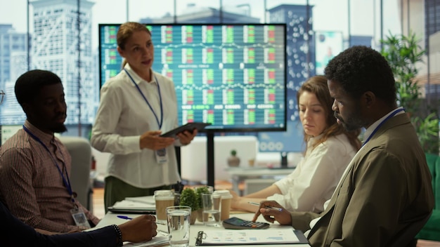 A diverse group of people discussing investment strategies around a table, with charts and graphs displayed on a screen.