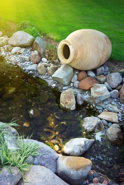 A close-up shot of a DIY water feature. The feature includes a collection of smooth river stones, a small recirculating pump, and aquatic plants, creating a tranquil and natural aesthetic.