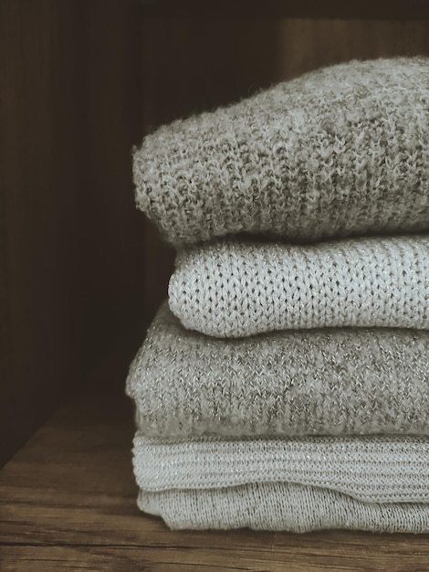 A close-up shot of a neatly folded stack of neutral-colored sweaters and t-shirts in a closet. The colors are muted and classic, including gray, beige, white, and black. The focus is on the texture and quality of the fabrics.