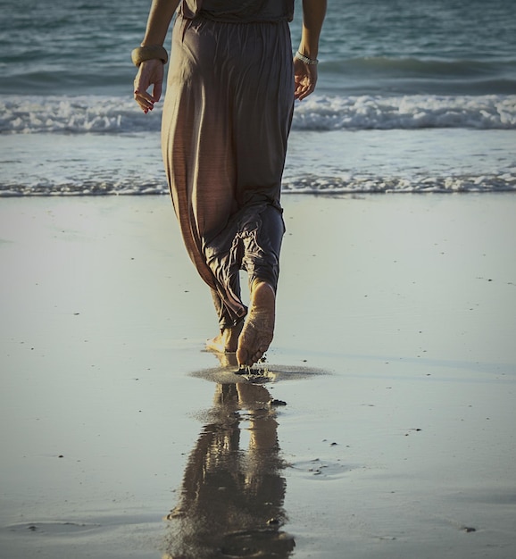 A person is walking barefoot on a sandy beach, with the ocean waves gently lapping at the shore. The sky is clear and blue, and the sun is shining brightly. The scene conveys a sense of peace and tranquility, emphasizing the connection with nature.