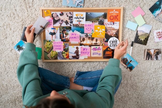 A close-up of a person's hands creating a vision board, carefully gluing images onto a corkboard. The images include pictures of dream homes, happy families, and exotic travel destinations.