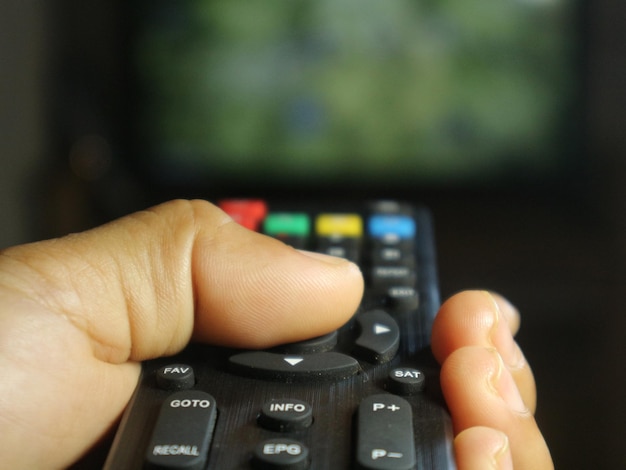A close-up shot of a person's hands holding a remote control, with a TV screen in the background displaying an intense scene from a popular drama series. The lighting is dim, creating a cozy and immersive viewing atmosphere.